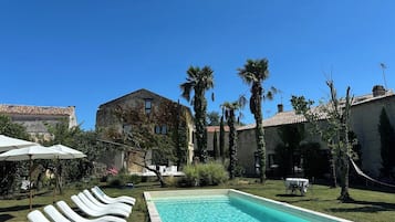 Una piscina con sillones de descanso, un edificio de piedra y palmeras al fondo.