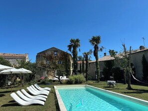 Una piscina con sillones de descanso, un edificio de piedra y palmeras al fondo.