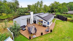 Ett modernt hus med en terrass, ett område för grillning och ett stort fönster med utsikt över en trädgård.