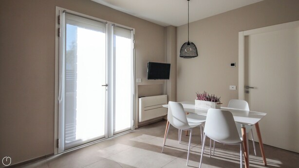 Un comedor moderno con una mesa y sillas blancas, un televisor de pantalla plana montado en la pared y un amplio ventanal corredizo que da a un espacio exterior.