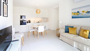 A modern living room with a sofa, a dining area with chairs, a kitchen with white cabinets, and a painting on the wall.