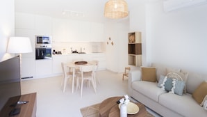 A modern kitchen with white cabinets, a dining area with a wooden table and chairs, a sofa with cushions, and a hanging light fixture.