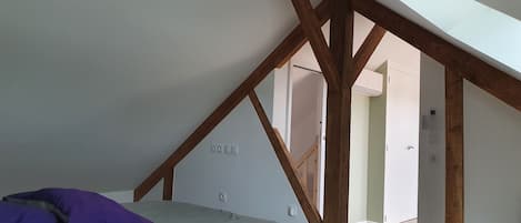 Un dormitorio con techo inclinado, vigas de madera a la vista, una cama con colchón de color claro y una almohada morada, y una silla en la esquina.