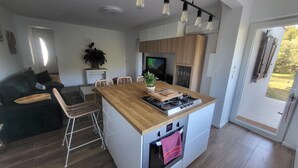 A modern kitchen with a central island, wooden cabinets, and a built-in oven. There is a dining area with a black sofa and wicker chairs. A potted plant is on the island, and a TV is mounted above the kitchen counter. - Villa Pour 7 Pers, 3 ch, Piscine Saint CYR SUR MER (Saint-Cyr-sur-Mer)