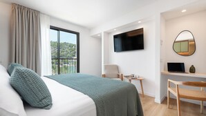 Habitació d'hotel moderna amb un llit gran, una taula de treball, una cadira, un televisor i un balcó amb vista a vegetació.
