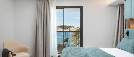 Een hotelkamer met een bed, een stoel, een balkon en uitzicht op zee en gebouwen.