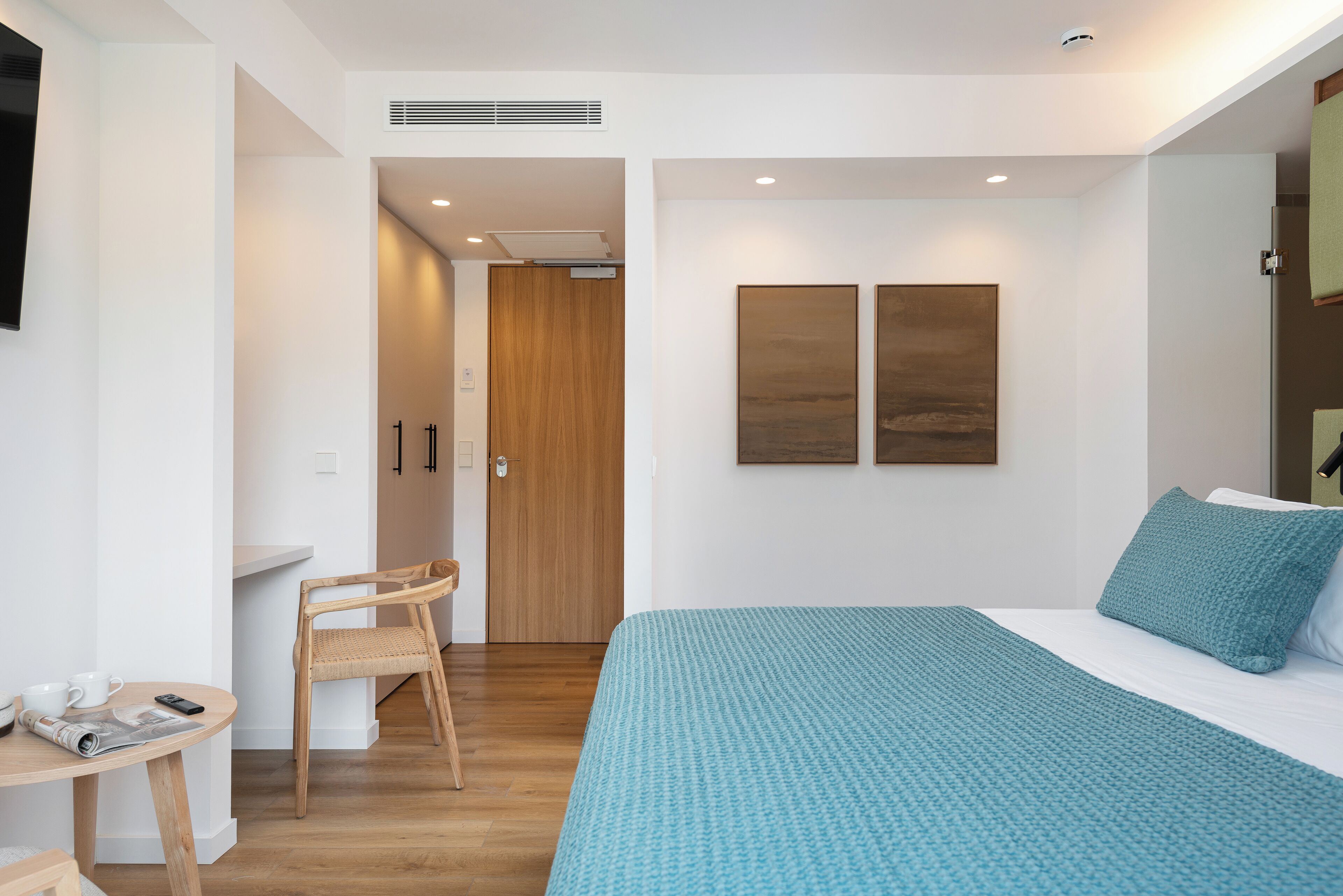 Habitación de hotel moderna con una cama grande, una silla de madera, una mesita pequeña y un escritorio. Hay una puerta que conduce a otra habitación y un televisor montado en la pared.