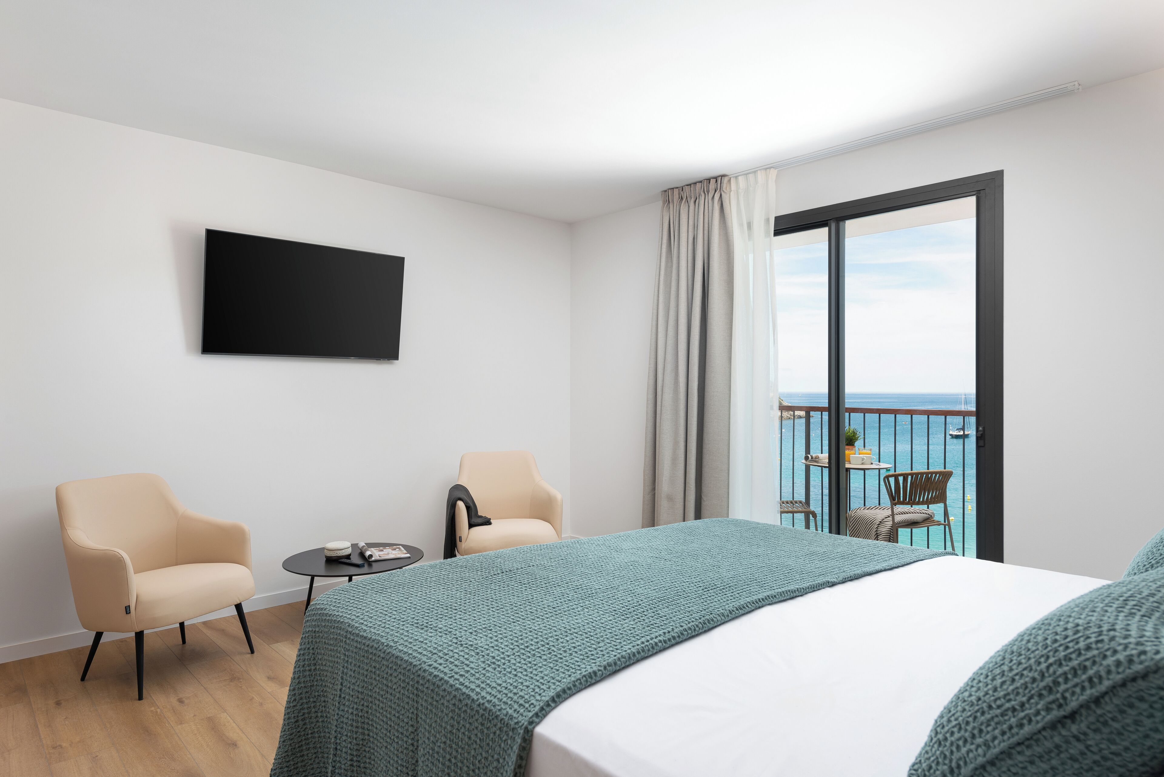 A hotel room with a bed, two armchairs, a small table, a TV, and a balcony with a view of the sea.