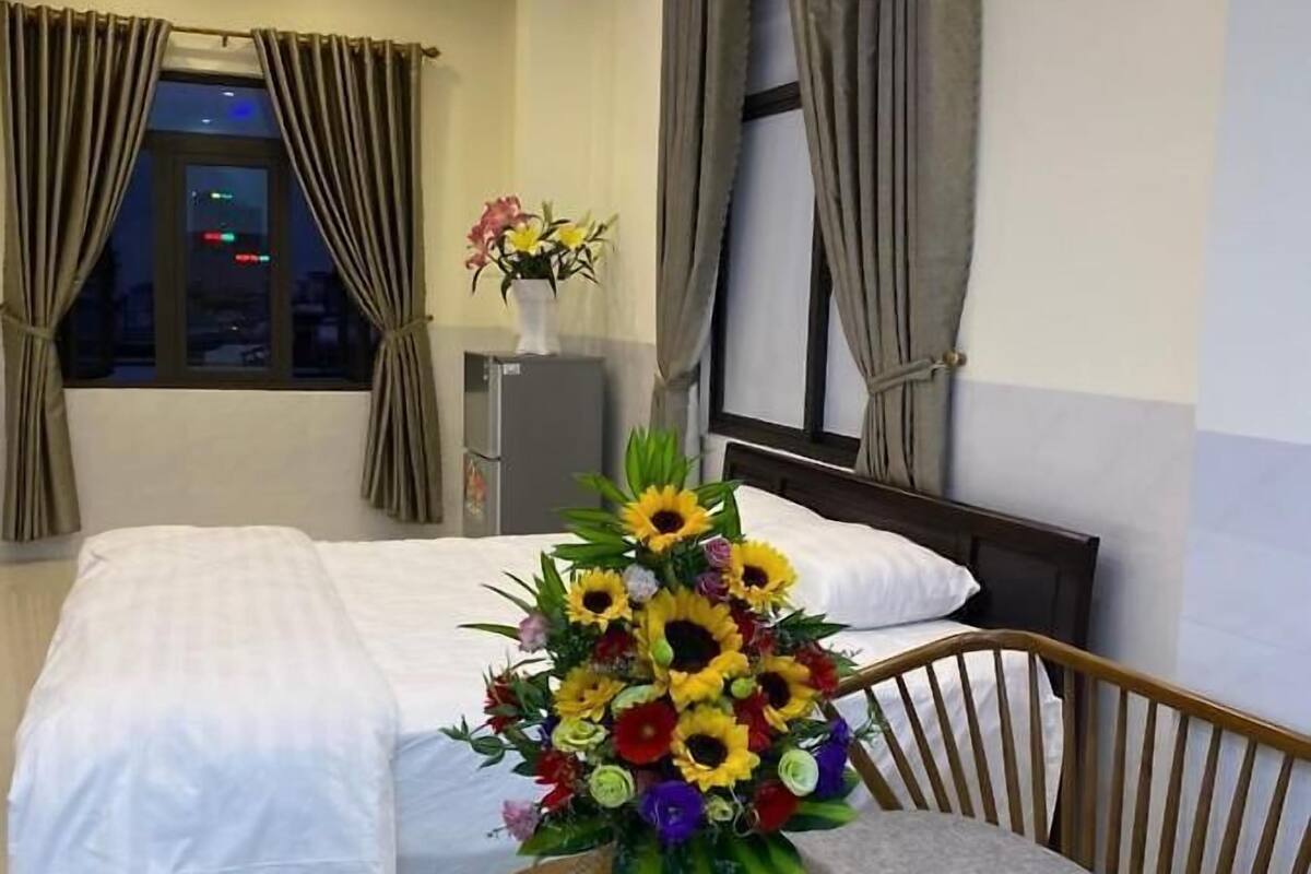 Una camera d'albergo con un letto, un tavolino con un vaso di fiori, una panca e una finestra con tende.