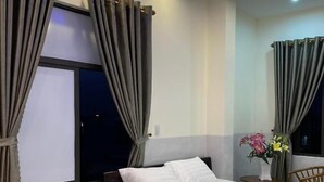 Een moderne slaapkamer met een groot bed, grijze gordijnen, een tafeltje met een bloevas en een raam met gordijnen.