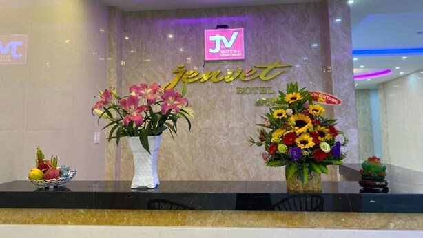 Een hotel receptiebalie met een televisiescherm waarop de naam van het hotel staat, een bord met de naam van het hotel en twee bloemstukken.