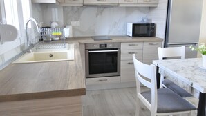 A modern kitchen with light wood cabinetry, stainless steel appliances, a marble countertop, and a dining area with white chairs. - Zennova 60 Nikiti Family Apt (Sithonia)