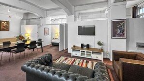 A modern living room with a black leather sofa, a dining area with a table and chairs, a TV mounted on the wall, and a small cabinet with speakers. - Maison Marvel l Enormous Loft Apt l Heart of City (Auckland)