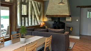 A rustic interior with exposed wooden beams, a stone fireplace, a dining area with a table and chairs, and a large window with curtains. - Stylish barn conversion  with a beautiful pool and stunning 4ha grounds. (Moustier)
