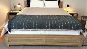 Una cama con estructura de madera, almohadas blancas y negras, y una manta rayada oscura. Hay dos mesitas de noche con lámparas y una planta pequeña.