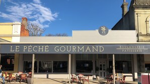 Fachada de un comercio llamado "LE PÉCHÉ GOURMAND" con mesas al aire libre y cielo despejado.