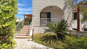 Un edificio moderno con una escalera de piedra que conduce a una entrada cubierta, rodeado de follaje y un cielo despejado.