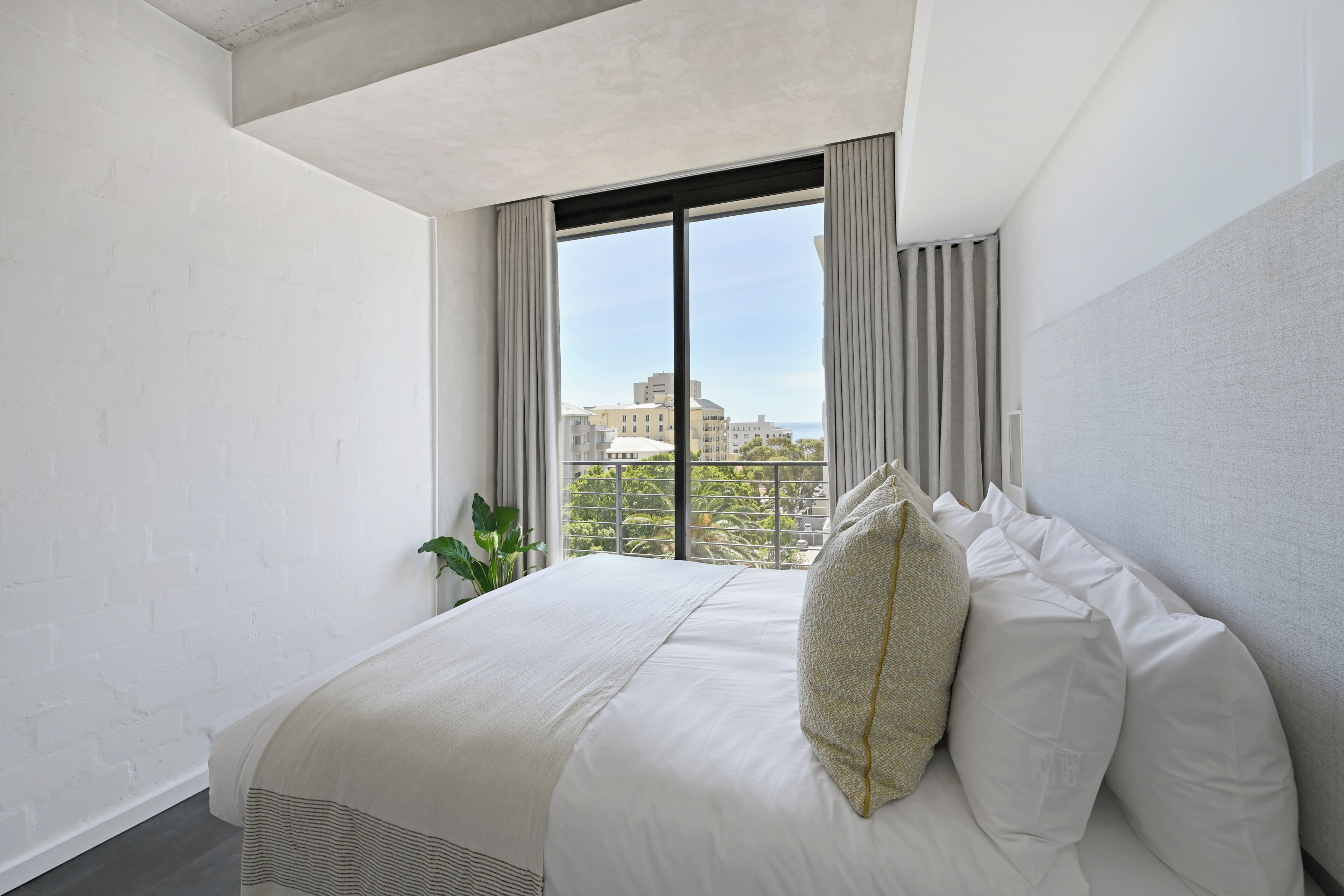 Une chambre d’hôtel moderne avec un grand lit, une vue sur les immeubles par la fenêtre et une plante en pot.