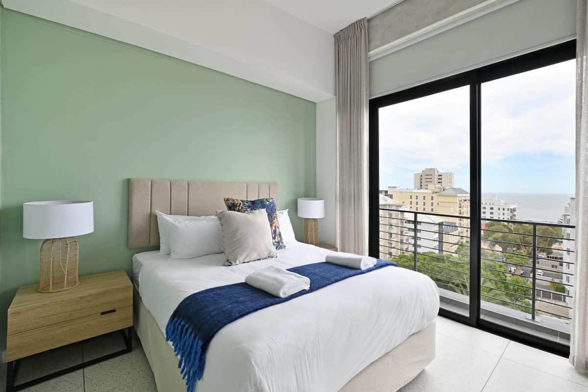 A hotel room with a large bed, two bedside tables with lamps, a view of the city through large windows, and a balcony.