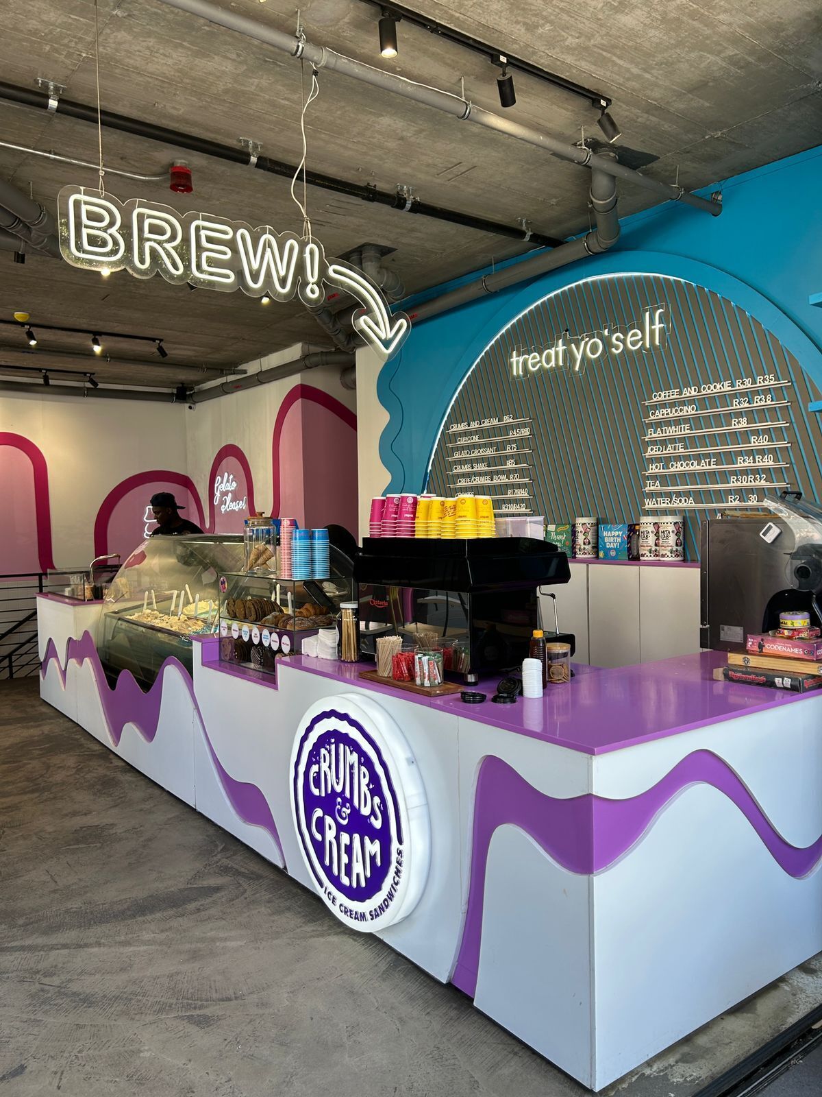 A counter with a neon sign reading 'BREW!' and a menu board listing various beverages and desserts.