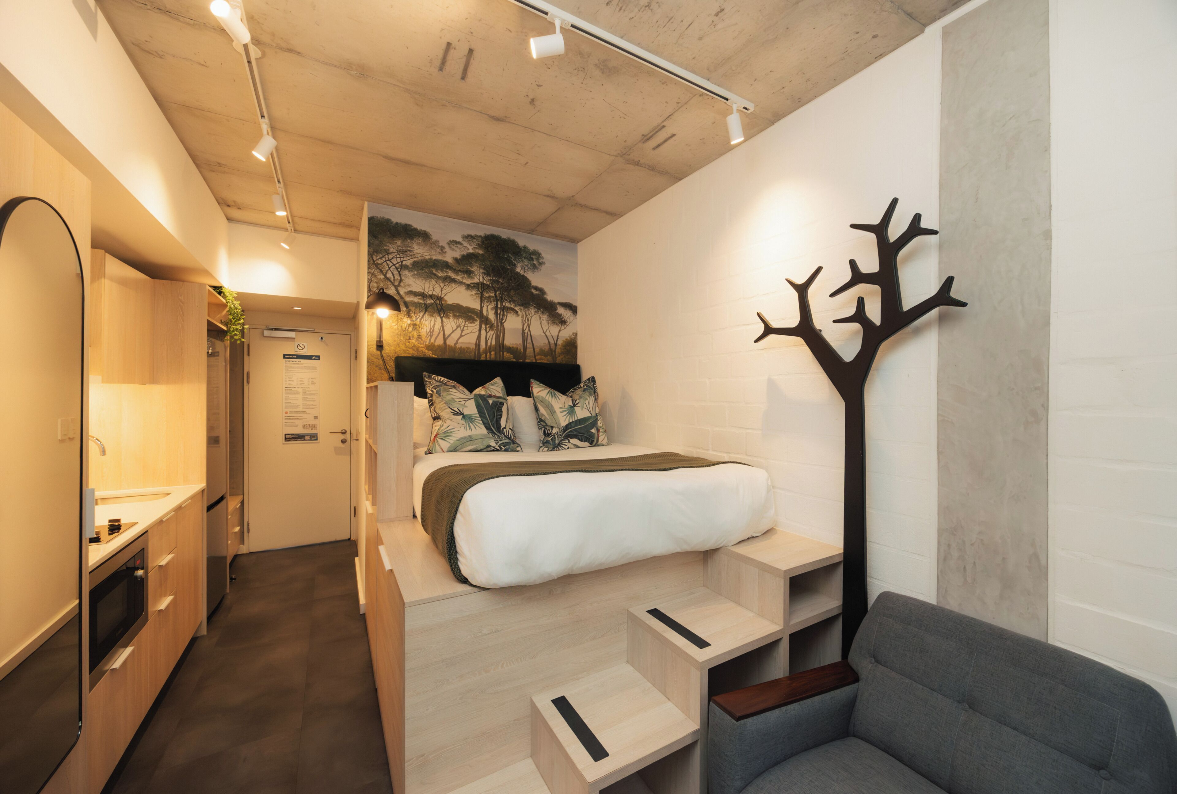 A compact hotel room with a bed, a seating area, a coat rack, and a wall-mounted artwork.
