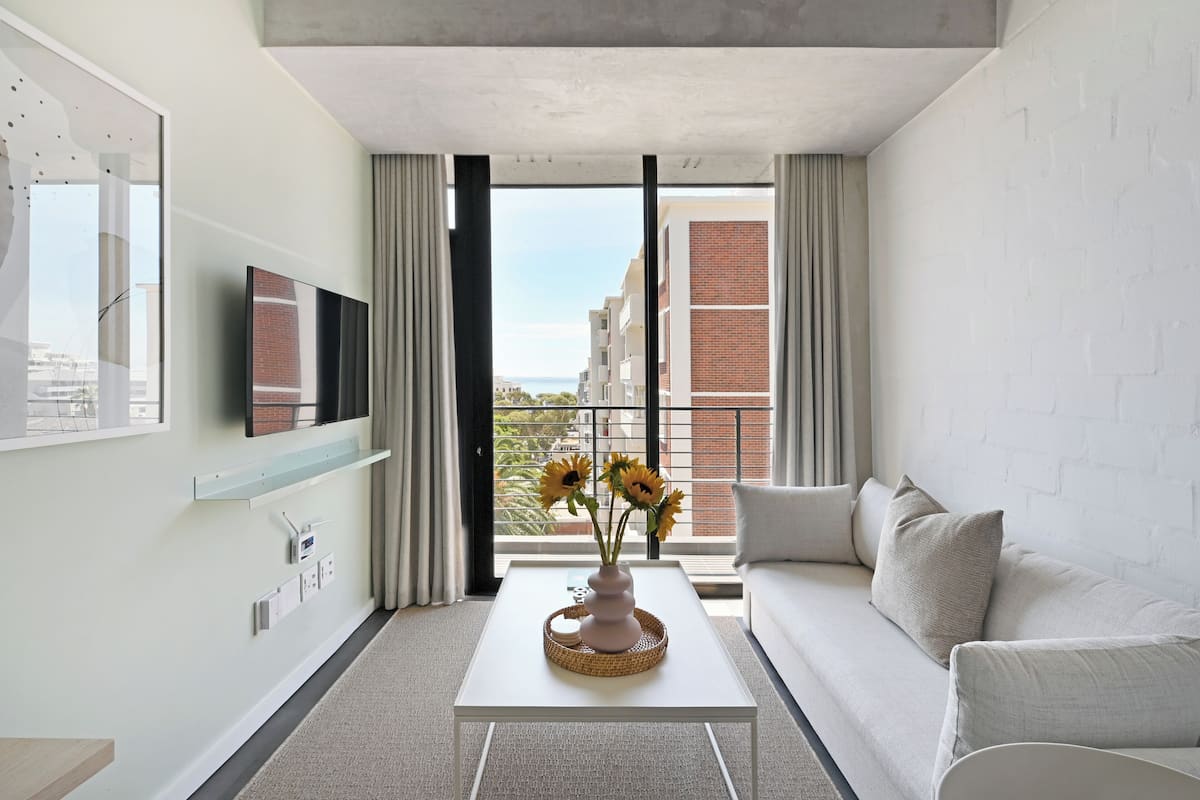 A modern living room with a white sofa, a coffee table with a vase of sunflowers, a flat-screen TV on the wall, and a large window with curtains.