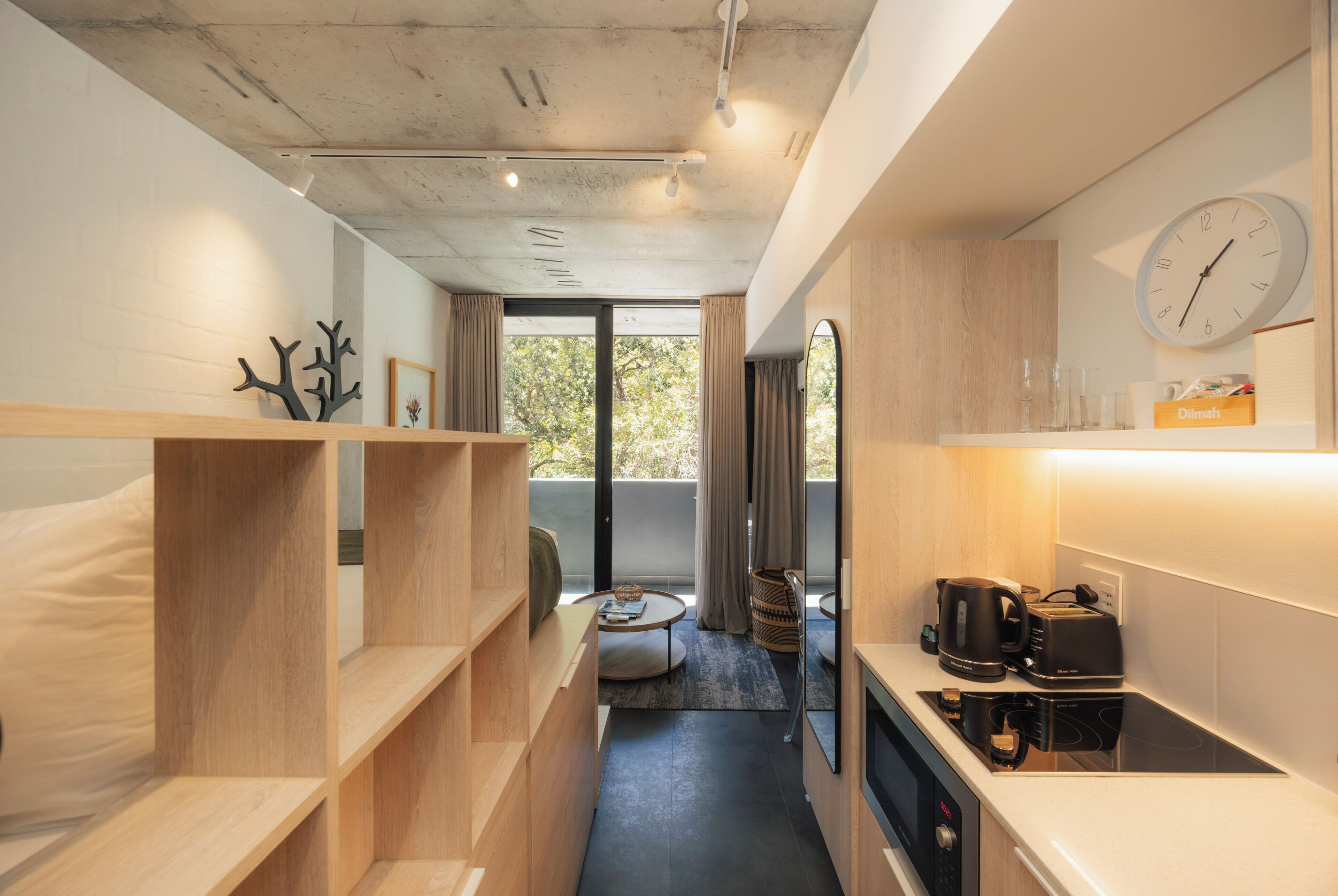 Une chambre d’hôtel compacte comprenant un coin cuisine, un lit, un petit espace salon et une vue dégagée sur l’extérieur grâce à une grande fenêtre.