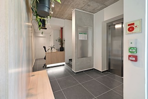 A modern lobby with a reception desk, a notice board, an elevator, and a fire alarm.