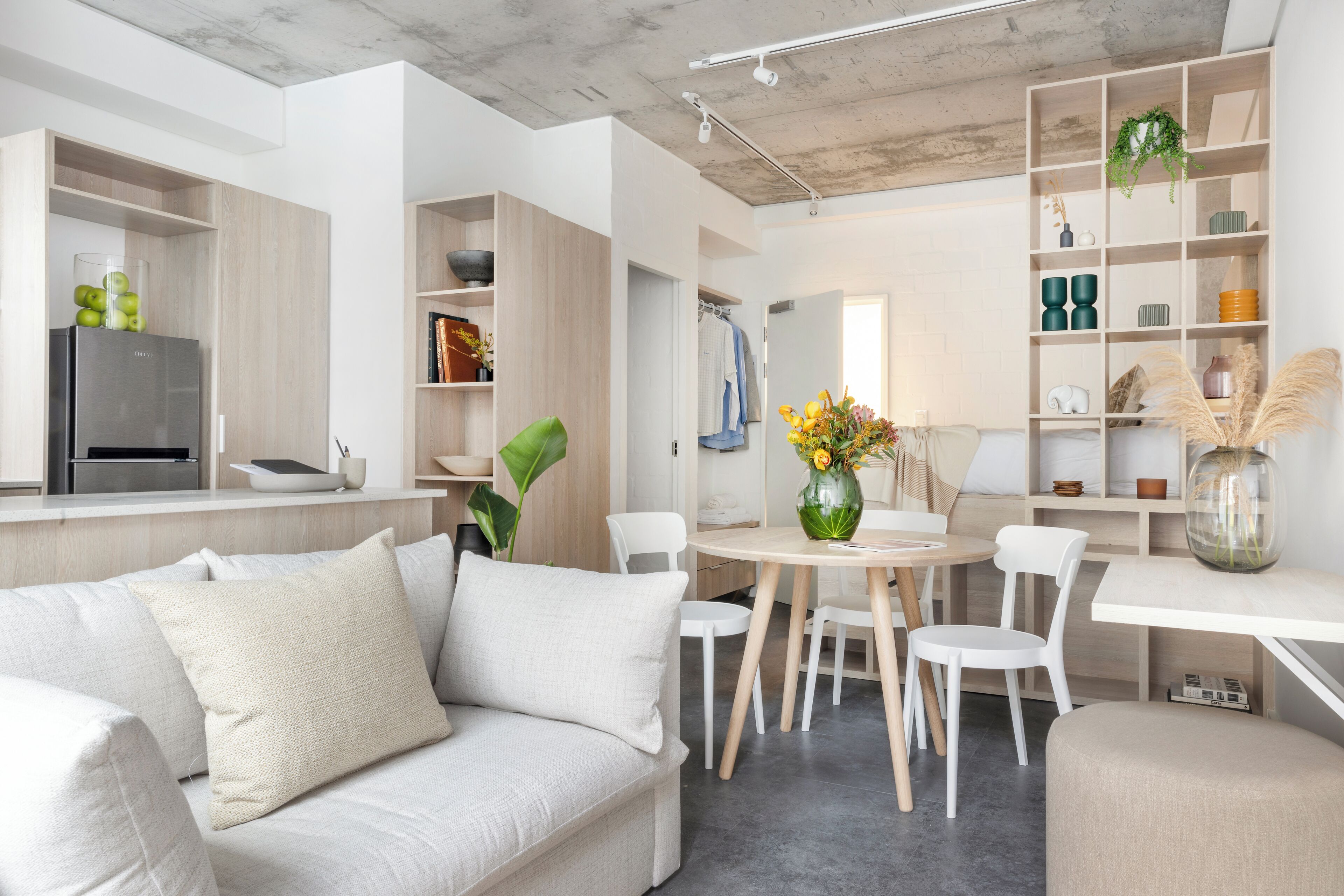 A modern living room with a sofa, dining table, chairs, and a kitchen area with a fridge and shelves.