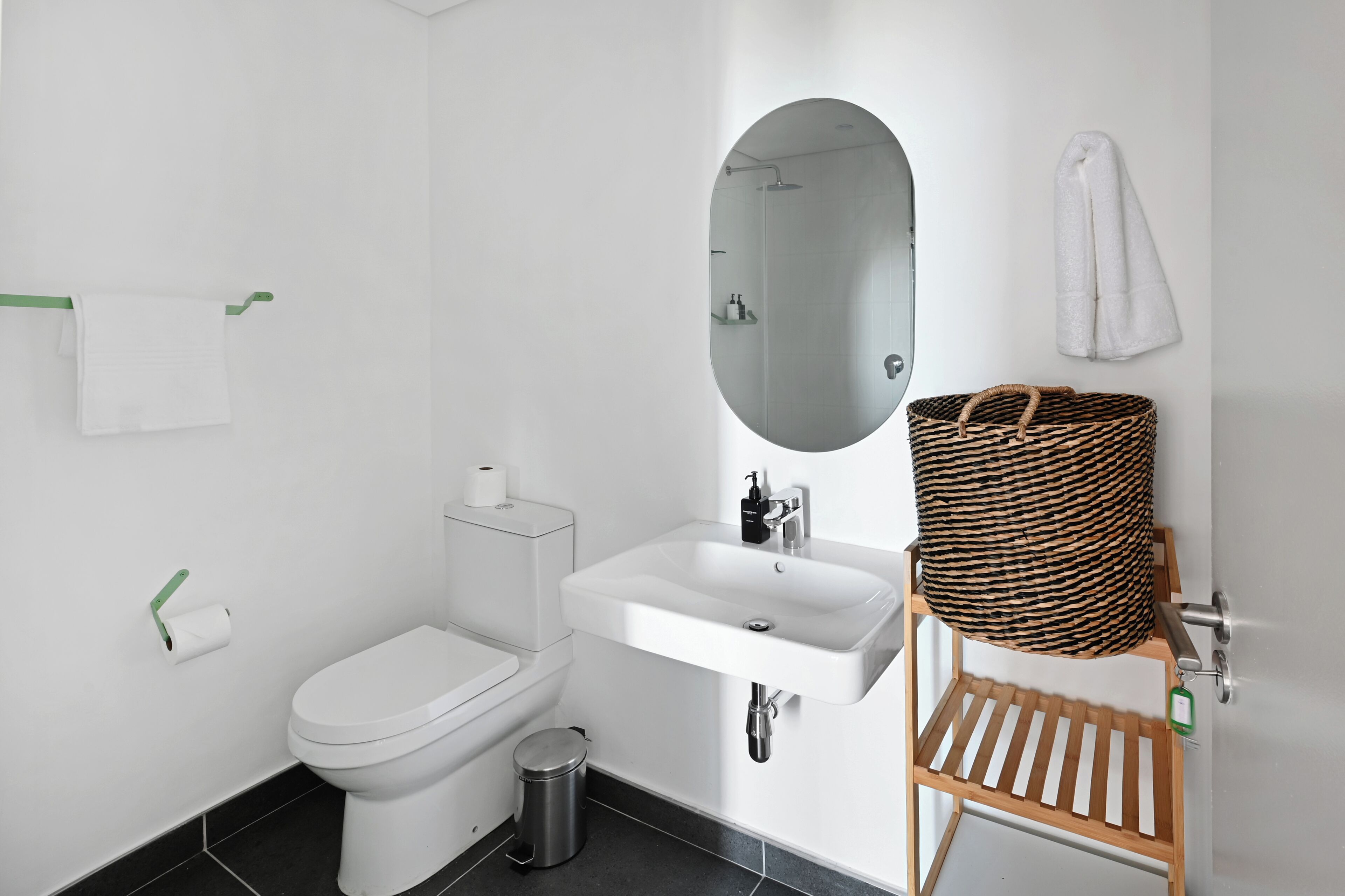 Ein modernes Badezimmer mit Toilette, Waschbecken und Spiegel. Ein Mülleimer und ein Korbwägelchen befinden sich auf einem Regal.