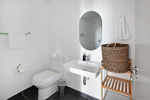 A modern bathroom with a toilet, sink, and mirror. There is a trash can and a wicker basket on a shelf.