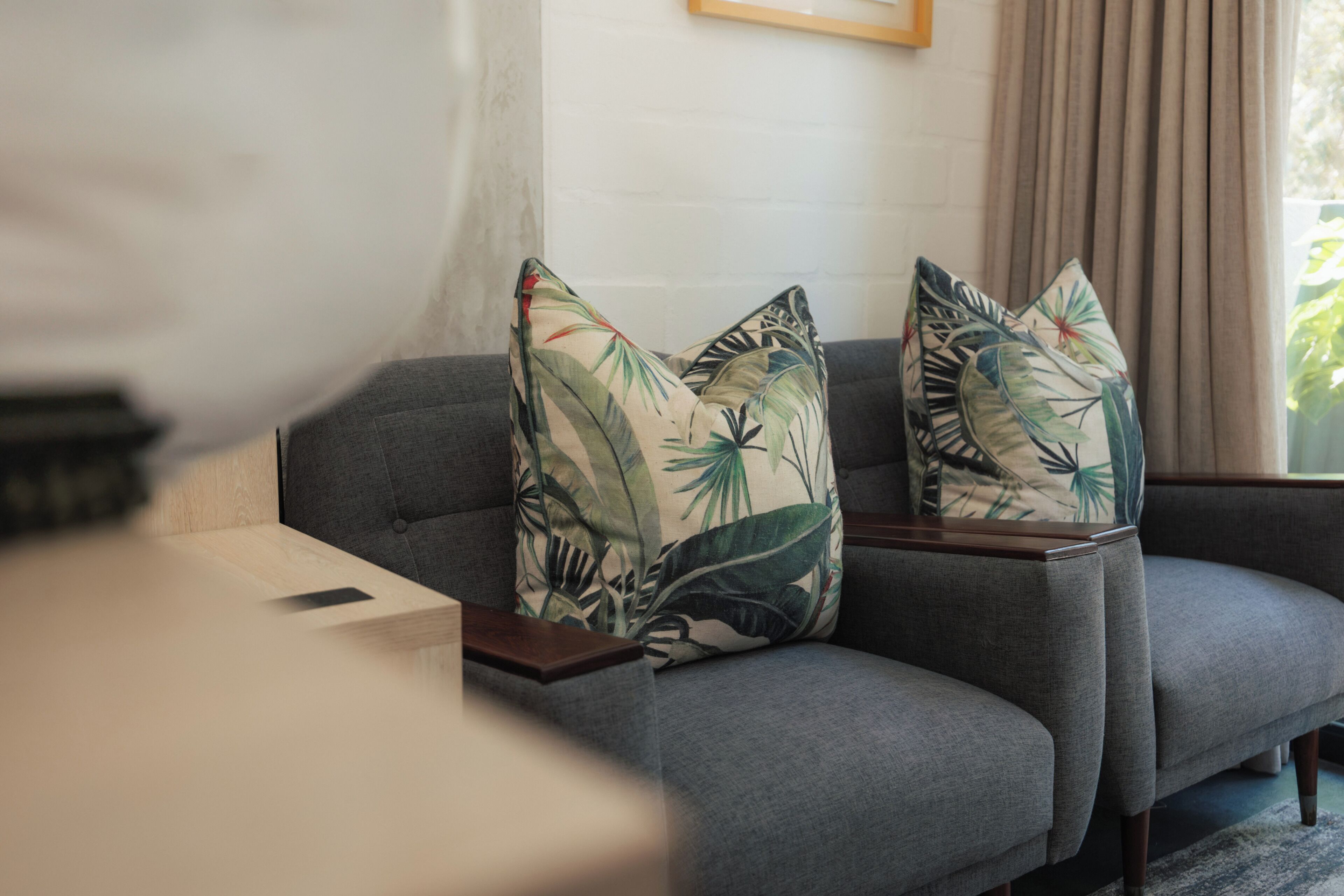 A grey sofa with tropical leaf-patterned cushions, a wooden side table, and a white wall with a picture frame.