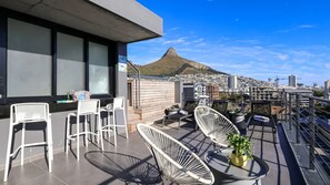 Eine Dachterrasse mit weißen Korbstühlen, einem kleinen Tisch und einem Blumentopf. Im Hintergrund ist eine Stadtansicht mit einem markanten Hügel zu sehen.