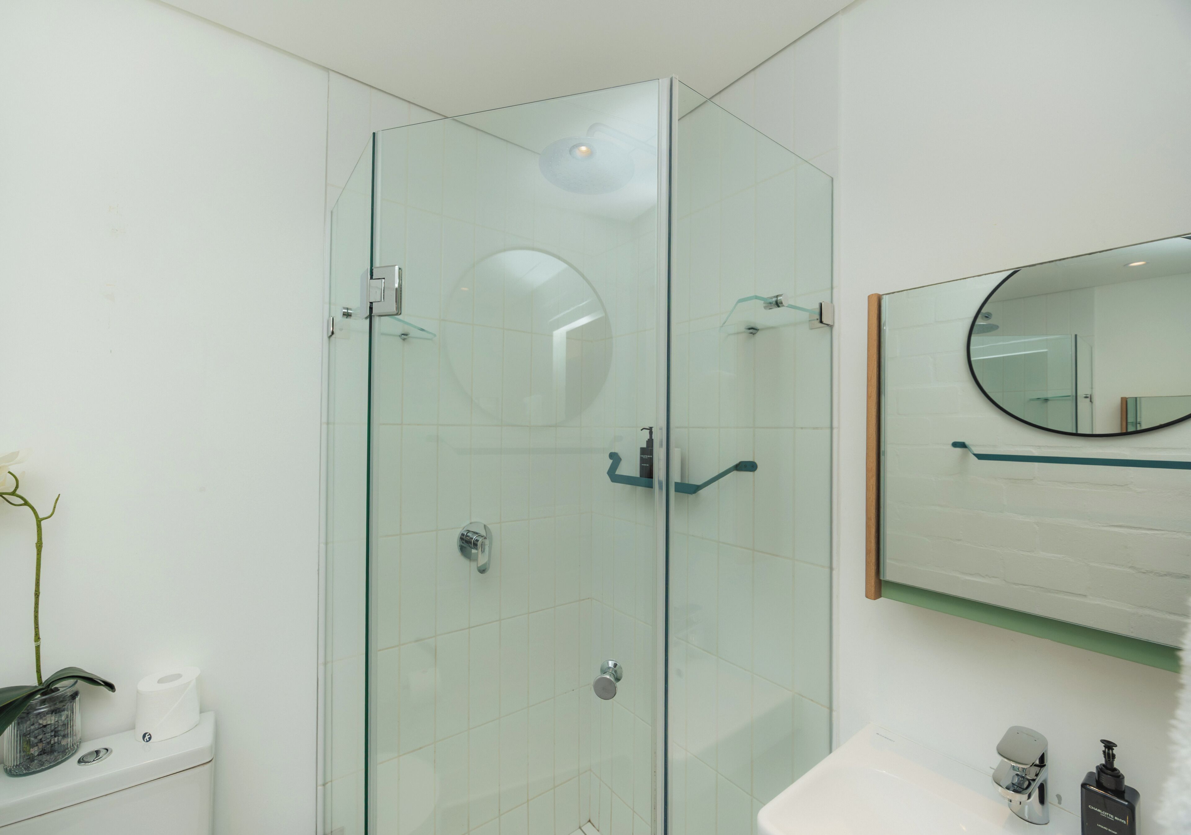 Un baño moderno con ducha envuelta en vidrio, un lavamanos blanco y un espejo con marco redondo.