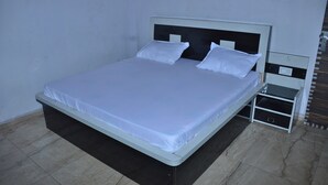 A neatly made bed with white linens and pillows, a bedside table with a lamp, and a power outlet on the wall. - Girdhar Sanwaria Palace (Danta Ramgarh)
