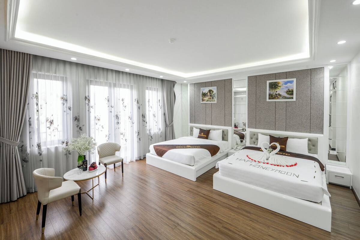 Una habitación de hotel moderna con dos camas, una zona de estar con una silla y una mesa, y ventanas amplias con cortinas.