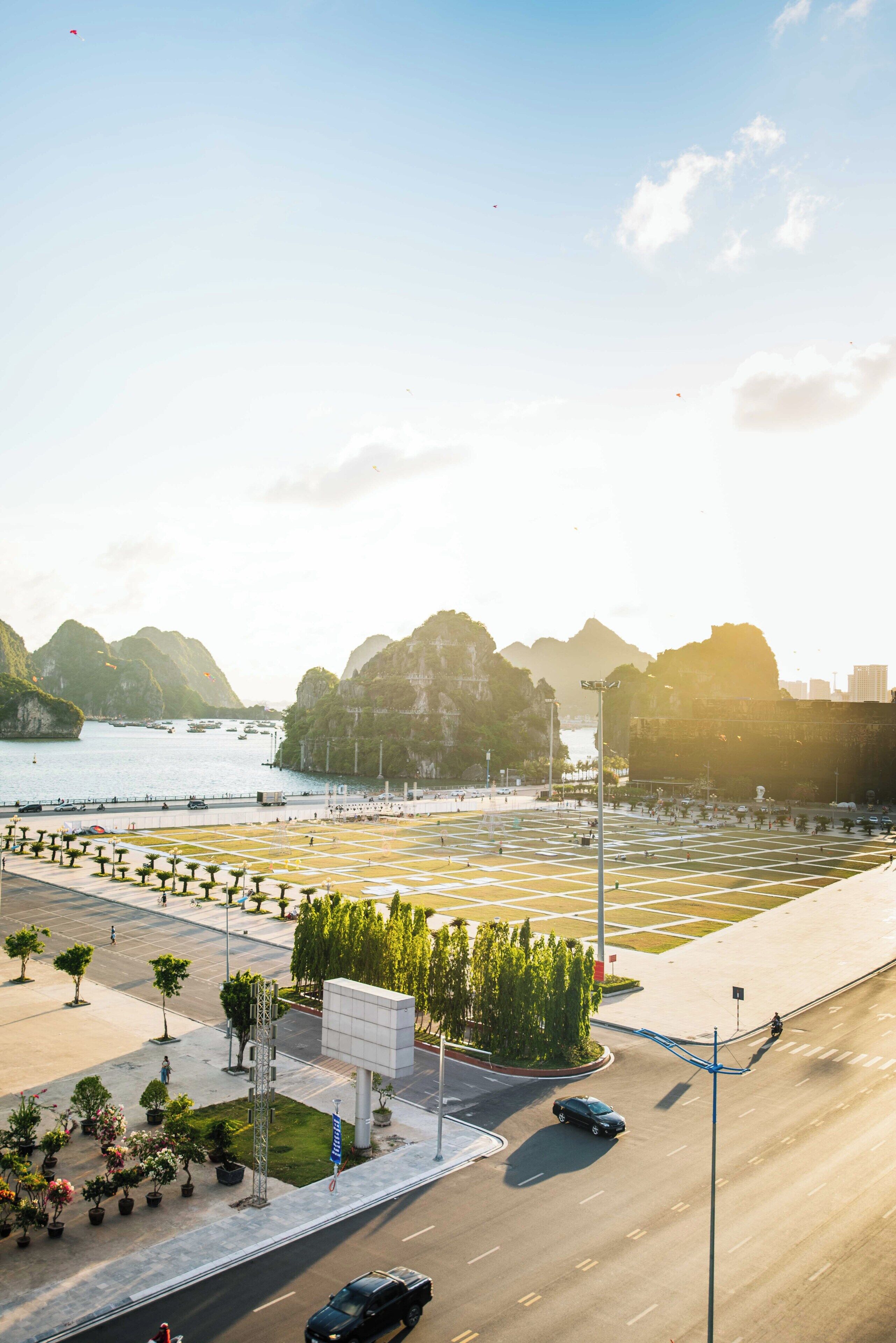 A wide road with a pedestrian crossing, a parking area, and a view of a body of water with boats.