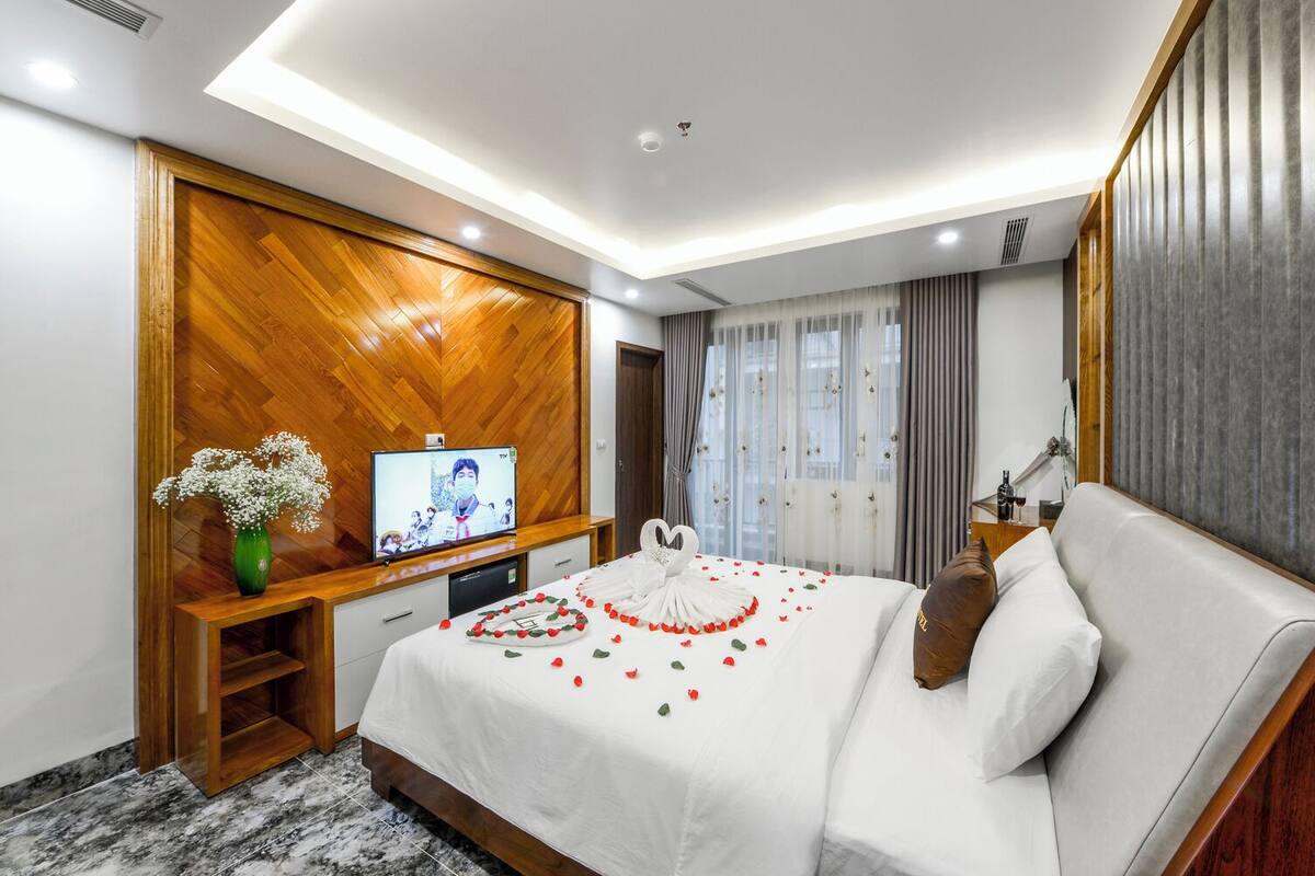 Habitación de hotel con una pared de acento de madera, un televisor, una cama con una colcha blanca y pétalos de flores rojas, y un florero con flores blancas.