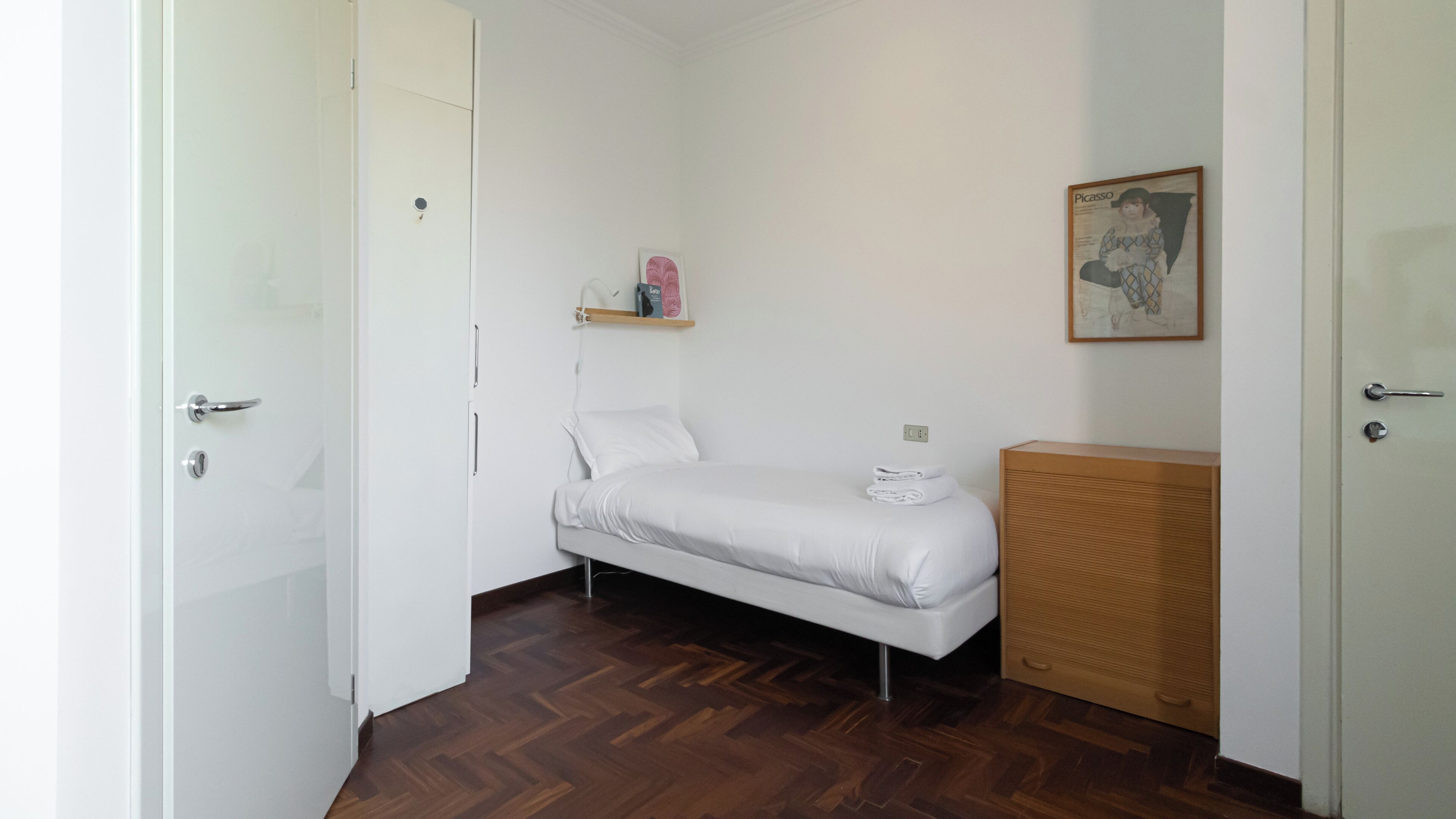 Ein kleines, ordentlich eingerichtetes Schlafzimmer mit einem Einzelbett, einem Holzschrank und einem Regal mit persönlichen Gegenständen.
