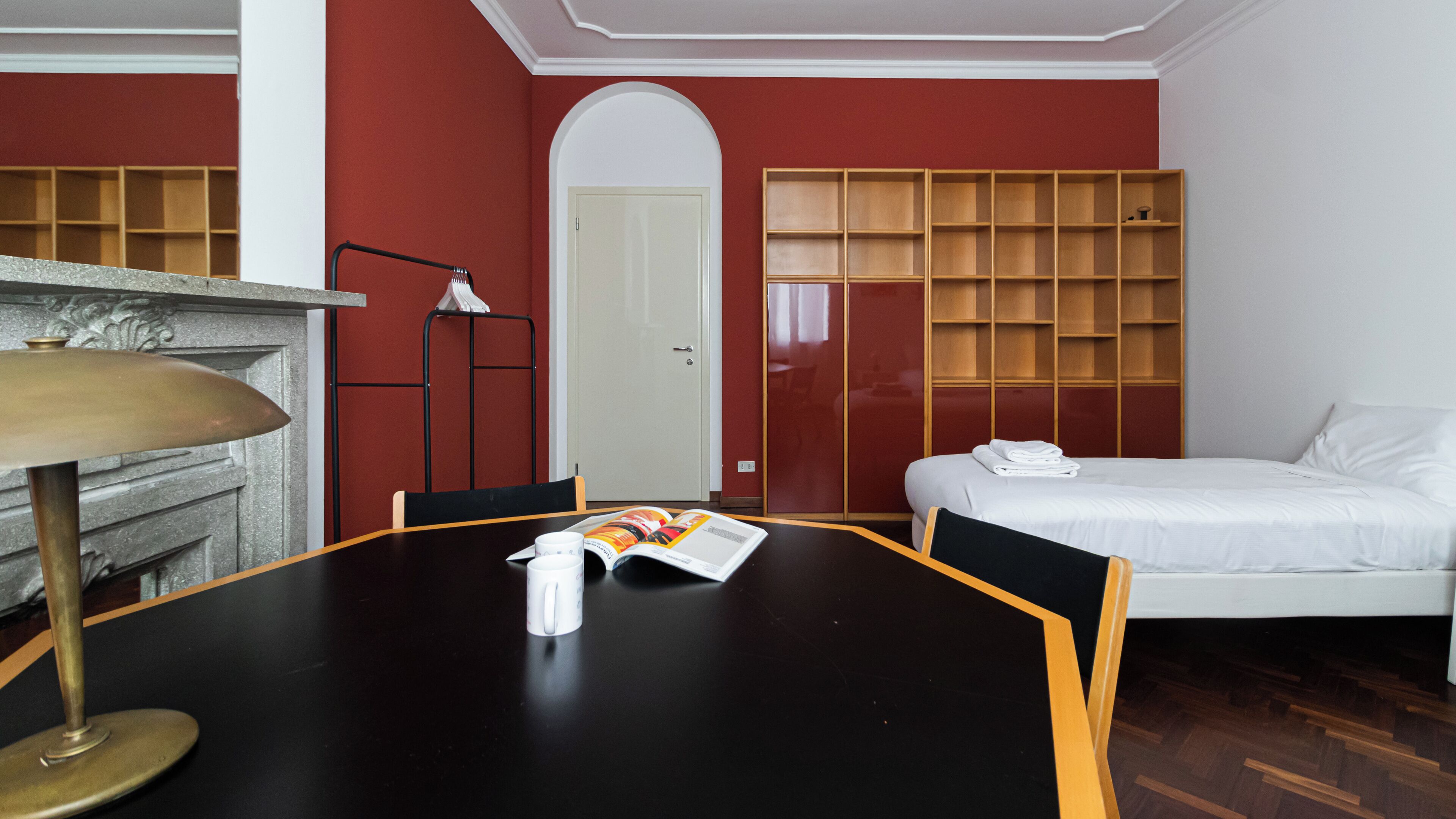 A hotel room with a black dining table, a white bed, a wooden bookshelf, and a white door.