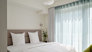 Un dormitorio moderno con un cabecero de madera, ropa de cama blanca y un ventanal con cortinas translúcidas.
