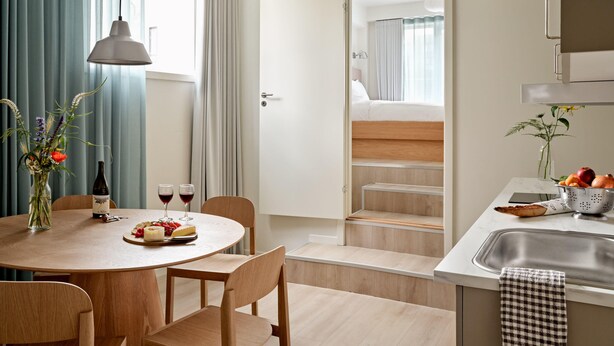 Una cocina moderna con comedor, muebles de madera, un lavamanos y vista a un dormitorio por una puerta abierta.