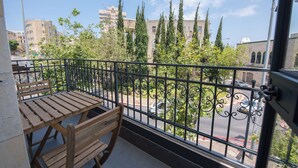 A balcony with a wooden table and chairs, a metal railing, and a view of a street with buildings and trees. - PORT CITY HAIFA -  Flea Market Luxury (Haifa)