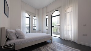 A modern bedroom with a large bed, white bedding, and a patterned rug. There are multiple windows with white curtains, and a balcony is visible through one window. - PORT CITY HAIFA -  Flea Market Luxury (Haifa)