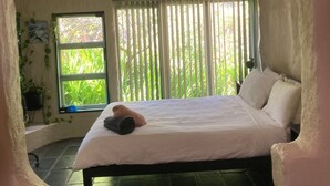 Ein Schlafzimmer mit einem Bett, einem Fenster mit vertikalen Jalousien, einer Topfpflanze und Blick auf Grünflächen.