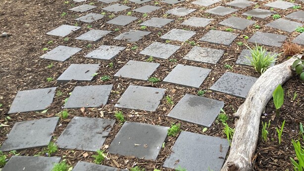 Ein gepflasterter Weg mit Steinplatten, umgeben von Mulch und kleinen Pflanzen.