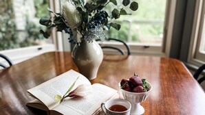 Een open boek, een kop thee, een schaal aardbeien en een vaas met bloemen op een houten tafel bij een raam.