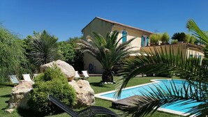 Zona de piscina con una piscina azul cristalina, rodeada de césped cuidado, palmeras y un edificio grande al fondo.