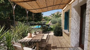 Un patio al aire libre con una mesa y sillas, una piscina al fondo y una zona cubierta con techo de tejido.