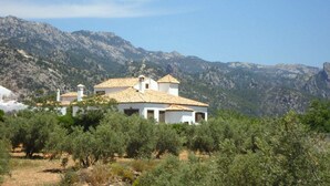Un bâtiment blanc, doté d’un toit en tuiles, entouré d’oliviers et avec des montagnes en arrière-plan.